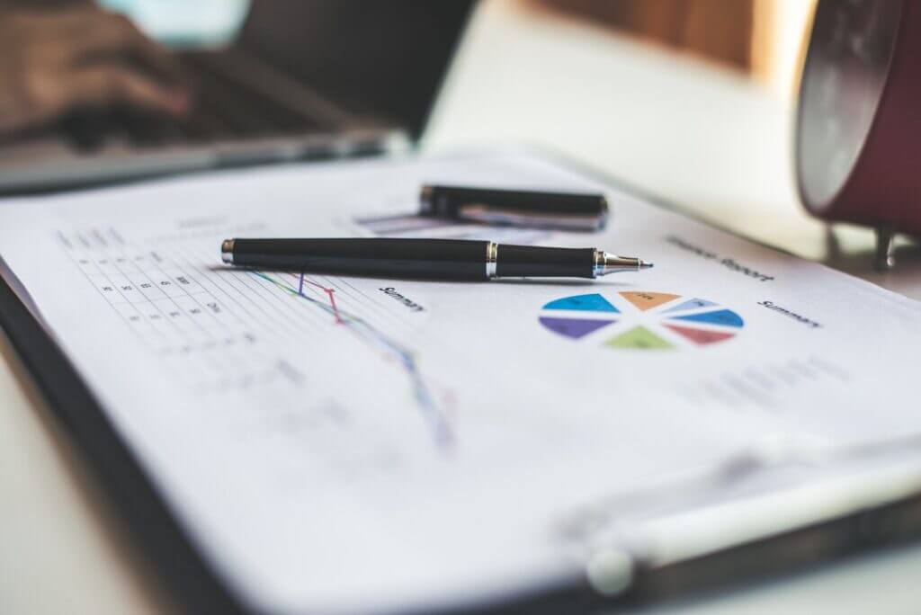 an uncapped pen rests on a financial report while a business person works on a laptop in the background